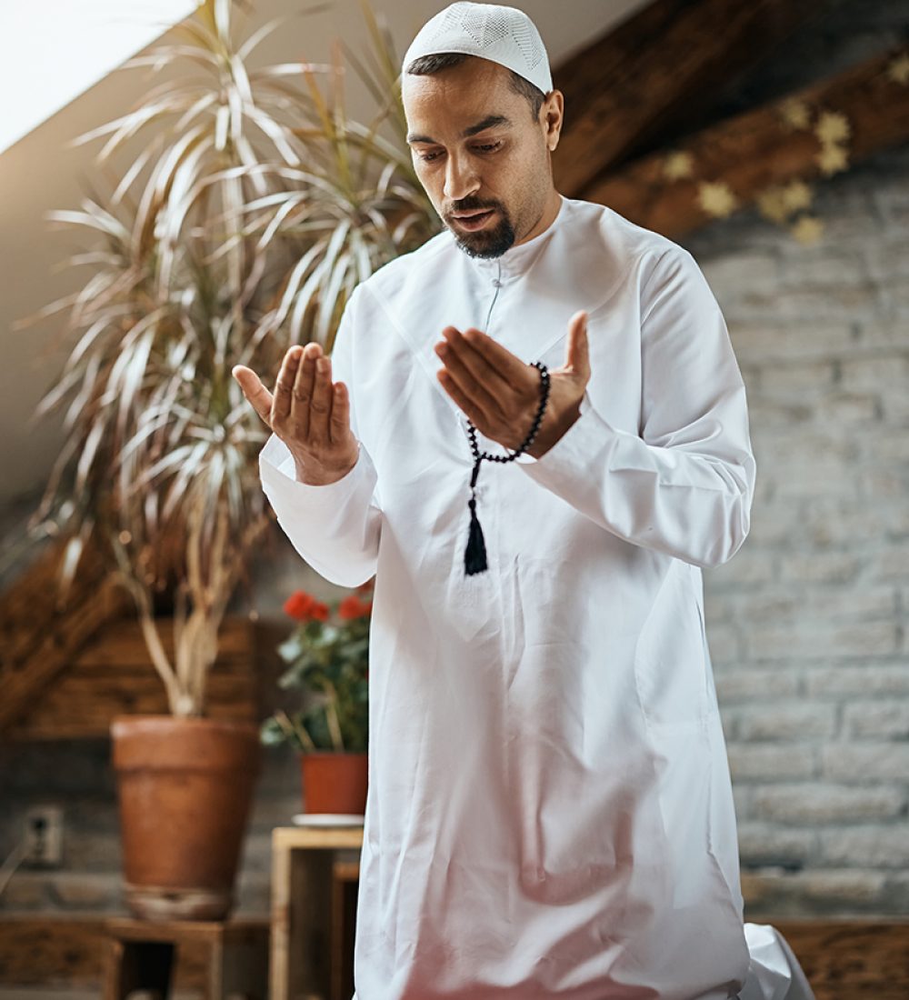 Religious Muslim man praying while kneeling on a prayer mat at home.